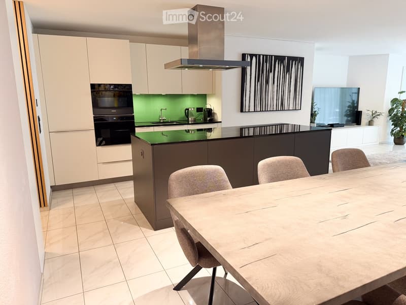 Modern kitchen with light-colored cabinets, a central island, and a dining area featuring a wooden table and chairs. Appliances include an oven, range hood, and coffee maker. A large screen is mounted on the wall, and a plant adds a touch of greenery.