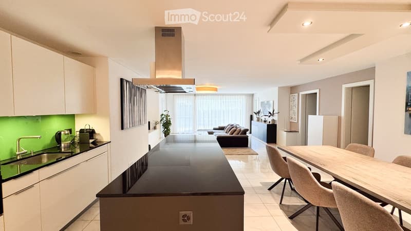 Modern kitchen with white cabinets, black countertop, and a green accent wall. Adjacent living area features a sofa, coffee table, and dining table with chairs.