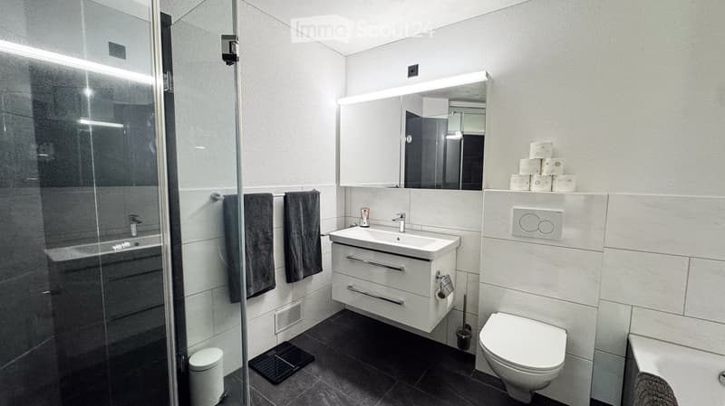 Modern bathroom with white tiles, a sink with a mirror cabinet, a toilet, a glass-enclosed shower, and neatly arranged towels and toilet paper.