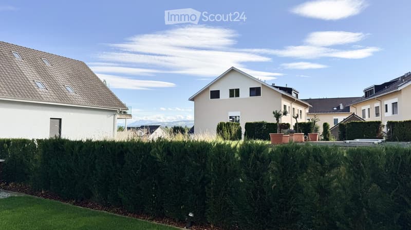 A well-maintained garden area with neatly trimmed hedges, potted plants, and a clear view of neighboring houses and a distant mountain range.