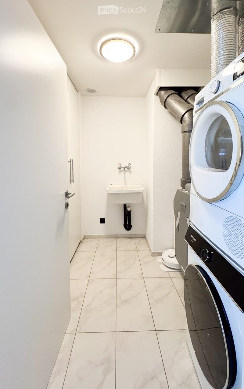 Modern bathroom with white tiled floor, sink, faucet, washing machine, and dryer. Features a skylight and sleek design.