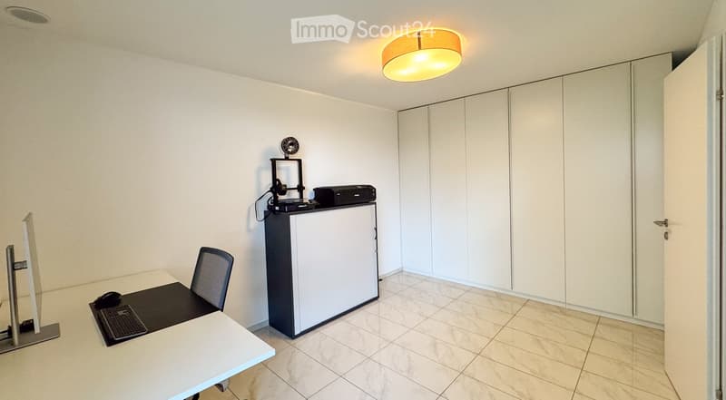 A modern, minimalist office space with a white desk, ergonomic chair, large screen, keyboard, mouse, small refrigerator, and printer on top. The room features white walls, tiled flooring, and built-in white cabinetry.