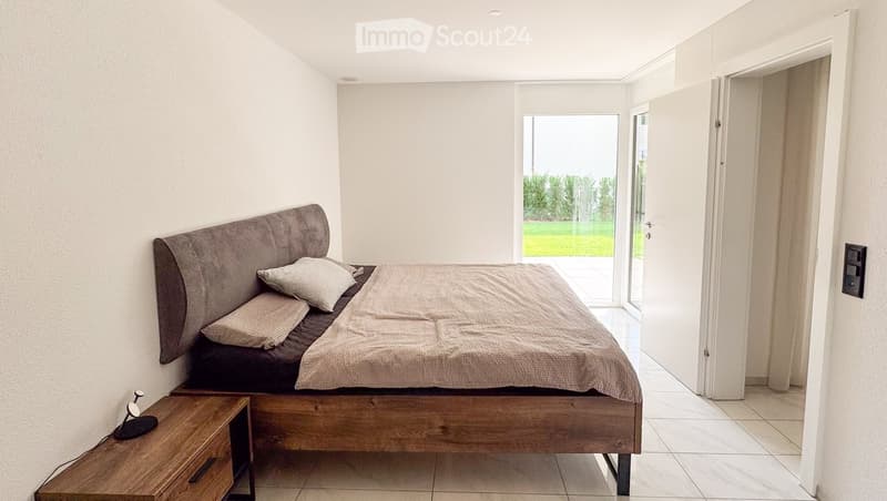 Modern bedroom with a large bed, wooden nightstand, and a lamp. The room features white walls, tiled flooring, and large windows with a view of a green outdoor area.