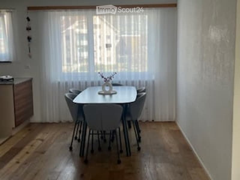 Dining room with a white table, 6 chairs, white curtains, wooden floor, a cabinet, and a vase with flowers on the table