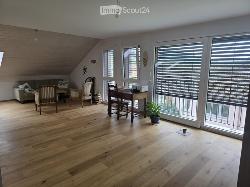 Bright, open room with wooden flooring, large windows, and a balcony. Features a sofa, chairs, a small table with a printer, and a potted plant.
