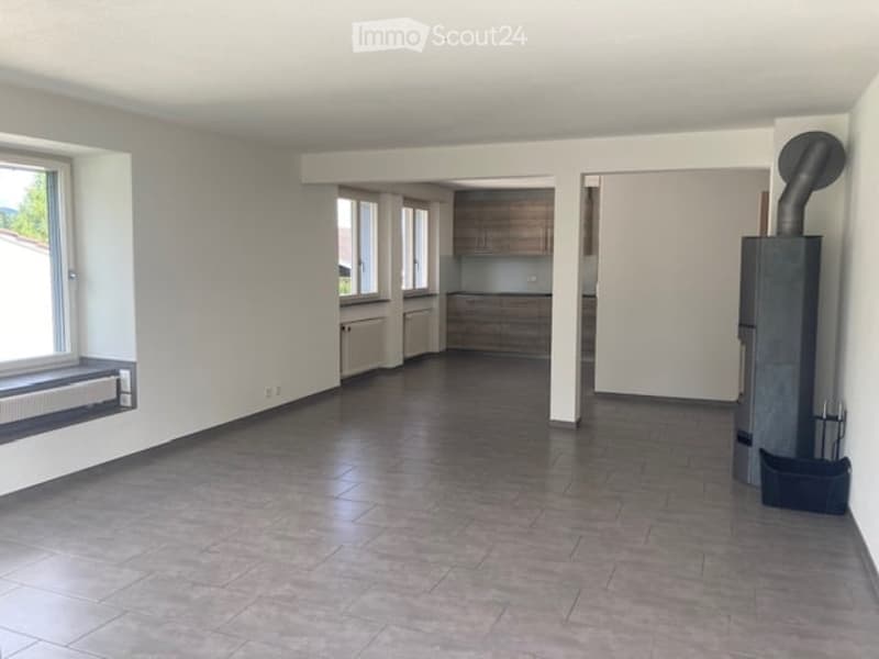 Empty room with tiled floor, white walls, large windows, and a small kitchen area visible in the background.