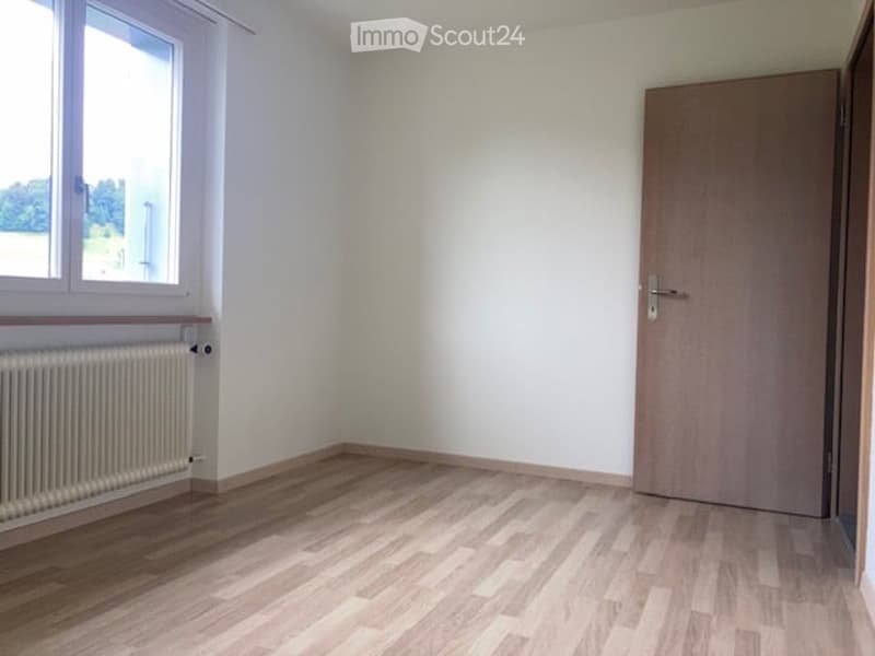 Empty room with light wooden flooring, white walls, a single window with white frame, and a closed wooden door.
