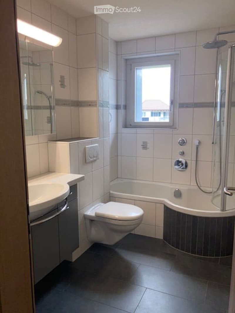 Modern bathroom with white tiles, a bathtub with a glass shower enclosure, a toilet, a sink with a mirror, and a window providing natural light.