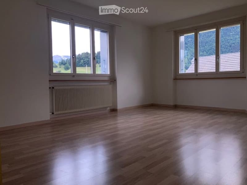 Empty room with wooden flooring, white walls, and large windows offering a view of greenery and mountains.