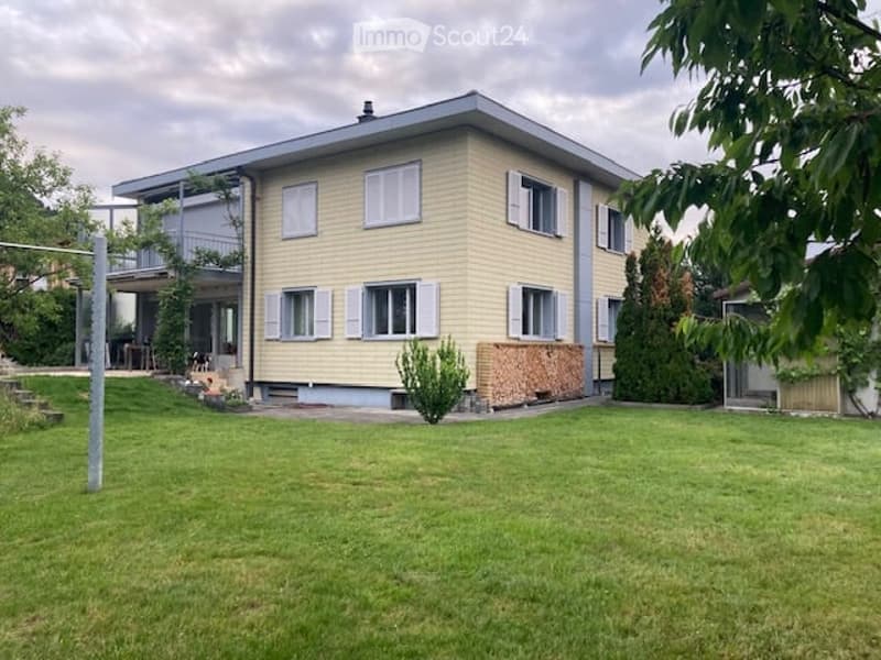 Two-story house with light yellow siding, white-framed windows, and a flat roof. The house features a covered balcony on the upper level and a well-maintained lawn in the front yard.