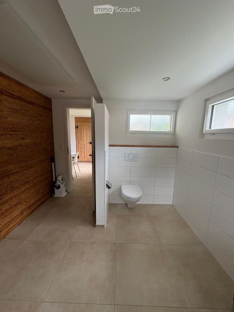 Modern bathroom with white tiled walls, beige tiled floor, and a toilet. The room has a wooden accent wall and a window above the toilet.
