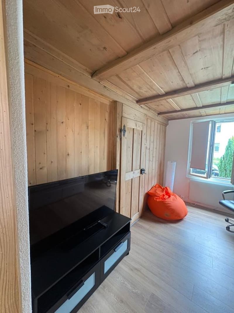 A modern living room with wooden walls and ceiling, featuring a large flat-screen TV on a black stand, an orange bean bag chair, and a black office chair near a window.