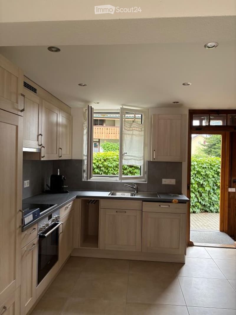 Modern kitchen with light wooden cabinets, black countertops, and stainless steel appliances. Features a window with a view of greenery outside.