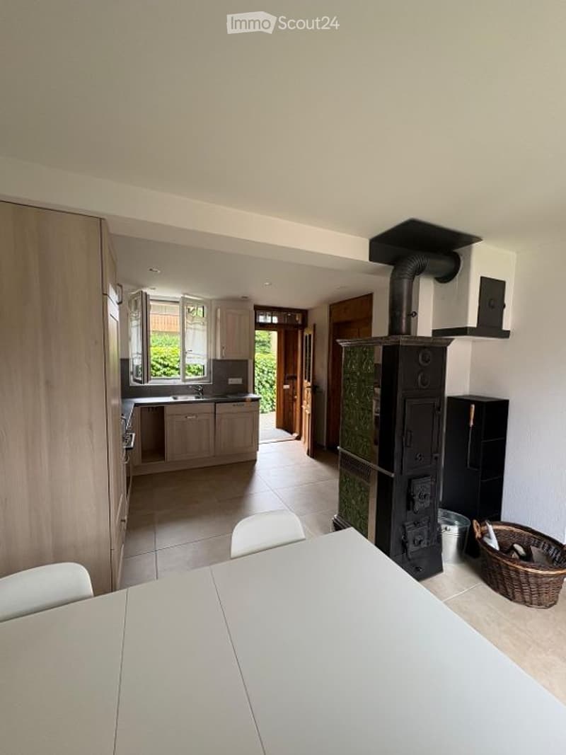 Bright, open-plan living and dining area with a modern wood stove, white dining table, chairs, and kitchen cabinets.