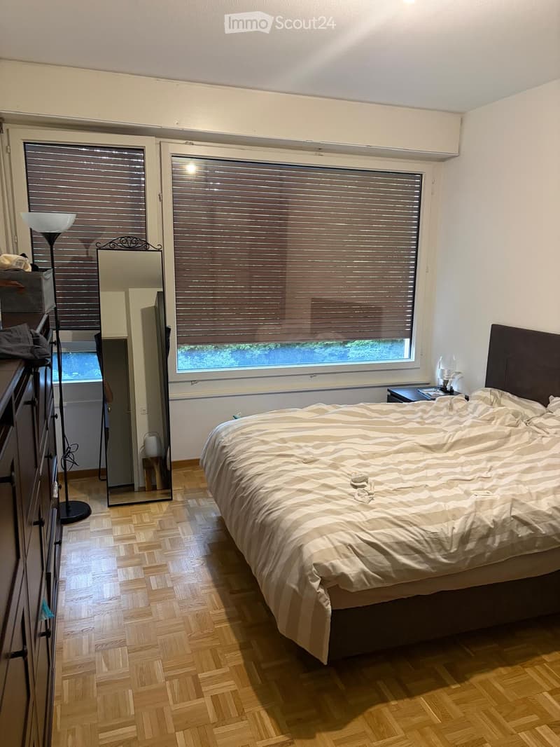 Cozy bedroom with wooden flooring, large window with blinds, neatly made bed, dresser, mirror, and floor lamp.