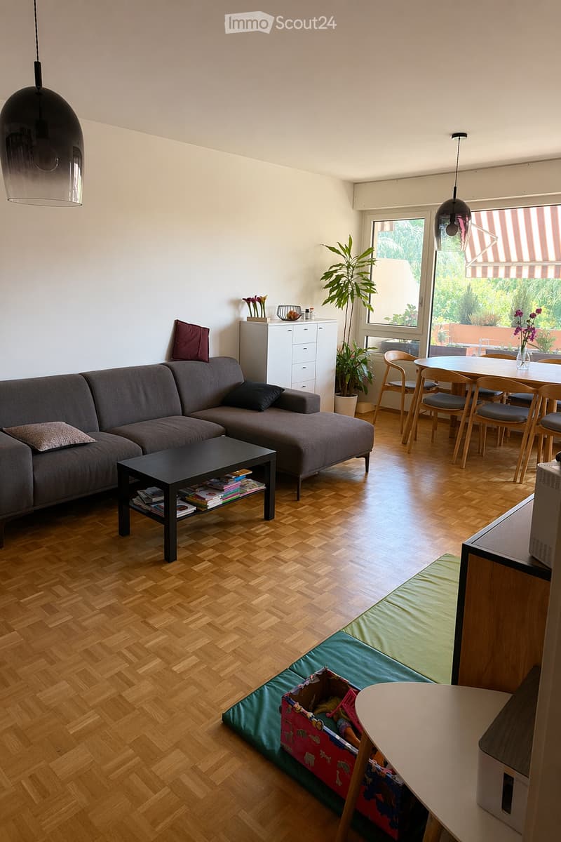 Living room with grey sectional sofa, black coffee table, dining area with chairs and table, pendant lights, and a small cabinet with decorative items.