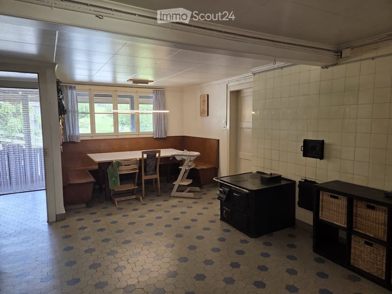 A cozy, well-lit room with a dining area and a small kitchen section. Features include a wooden bench seating, a table with chairs, a baby chair, a black stove, and a cabinet with wicker baskets.
