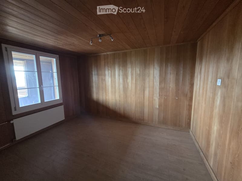 Empty room with wooden walls, wooden ceiling, and a large window with white frame. The floor is wooden, and there is a radiator below the window.