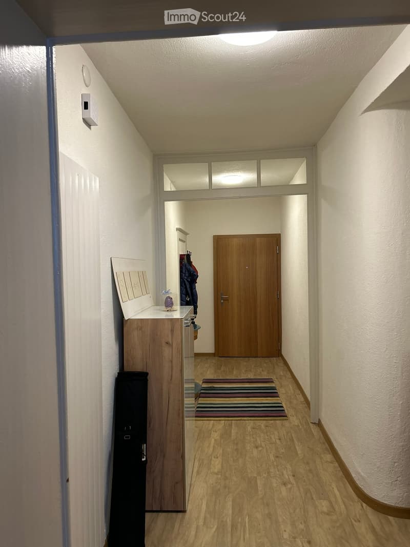 A narrow hallway with wooden flooring, white walls, and a small rug. A wooden door is visible at the end, and a small console table with a few items sits against the left wall.