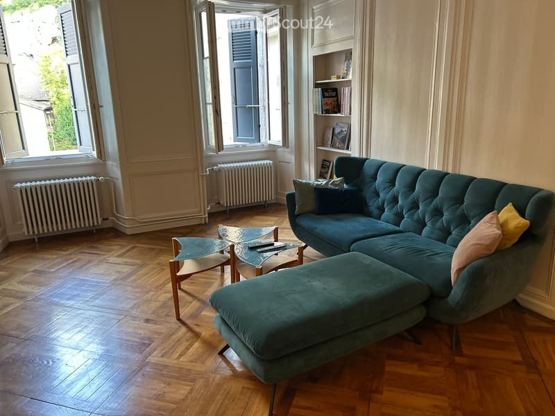 Living room with a green sectional sofa, ottoman, coffee tables, bookshelf, and radiators near the windows.