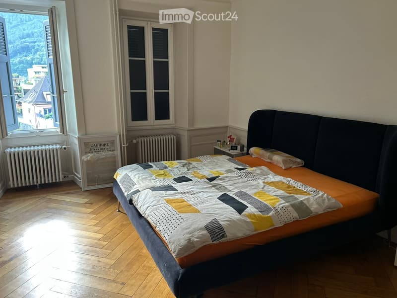 Bright bedroom with wooden flooring, a single bed with a colorful quilt, a nightstand, and a radiator near the window.