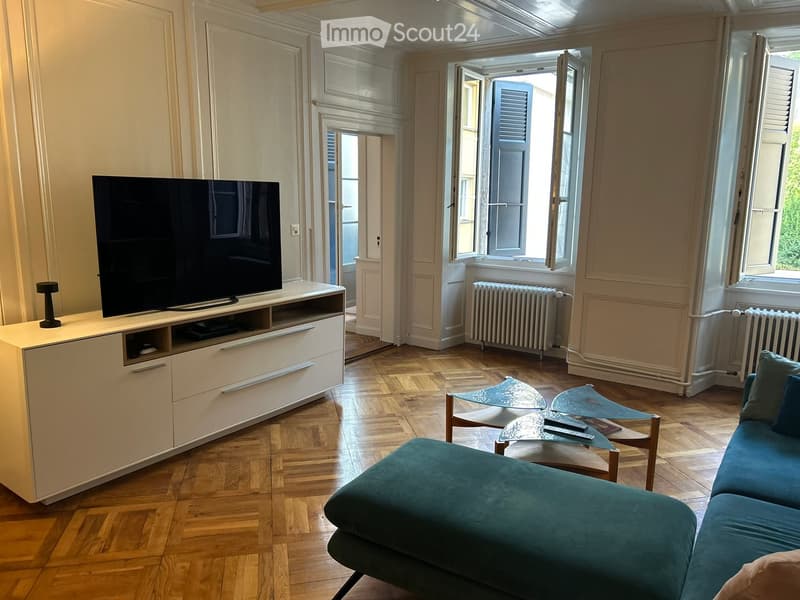Living room with wooden flooring, large windows, a teal sofa, a glass coffee table, a flat-screen TV on a white stand, and a table lamp.