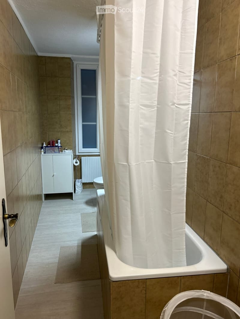 Modern bathroom with beige tiles, a bathtub, a white cabinet, a toilet, and a radiator. The space includes a shower curtain and a small shelf with toiletries.