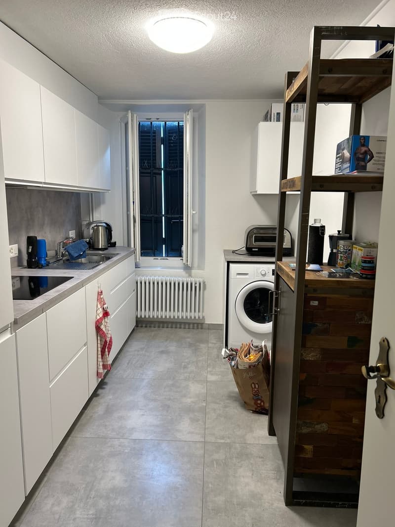 Modern kitchen with white cabinets, stainless steel sink, washing machine, and various appliances. Includes a wooden shelf with items and a small table with cups and bottles.