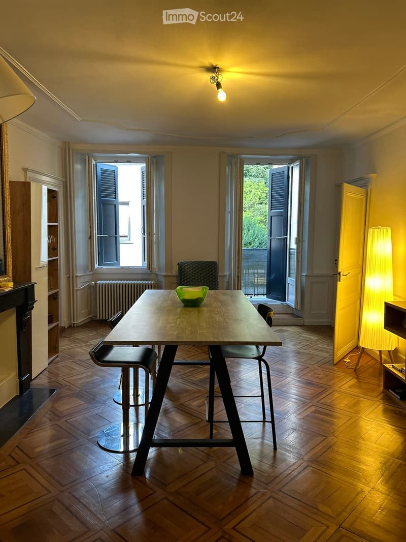 Dining area with wooden herringbone floor, modern table with four stools, green bowl on table, large windows with shutters, warm lighting from pendant lamp and floor lamp.