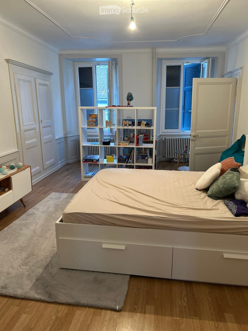 A well-lit bedroom with a large bed, a bookshelf filled with books and decorative items, a small table with a lamp, and a radiator near the window.