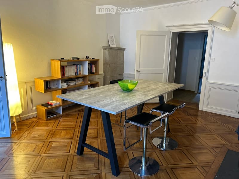 A modern, well-lit room with a large marble table, four black stools, a wooden shelf with books, and a green bowl on the table.