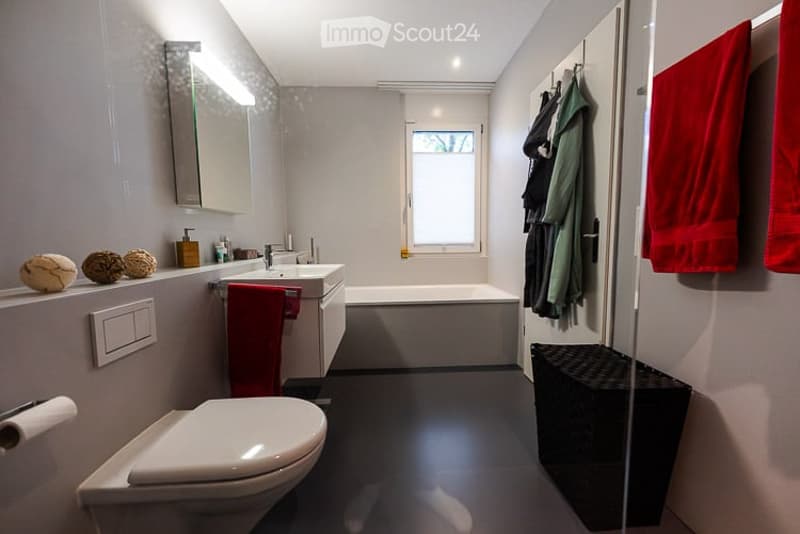 Modern bathroom with white walls, black floor, white toilet, sink, bathtub, mirror, and red towels. Decorative spheres on the windowsill and a light switch visible.