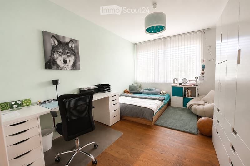 A modern, well-lit bedroom with a bed, desk, chair, and a small sofa. The room features a large window with sheer curtains, a hanging pendant light, and a picture of a wolf on the wall.