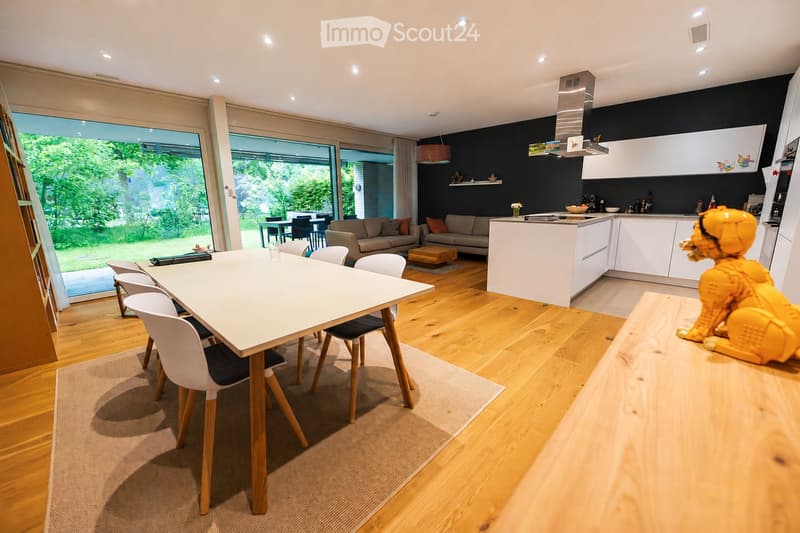 Open-plan living and dining area with a white dining table and chairs, a grey sofa, a coffee table, and a modern kitchen island with white cabinets and stainless steel appliances.