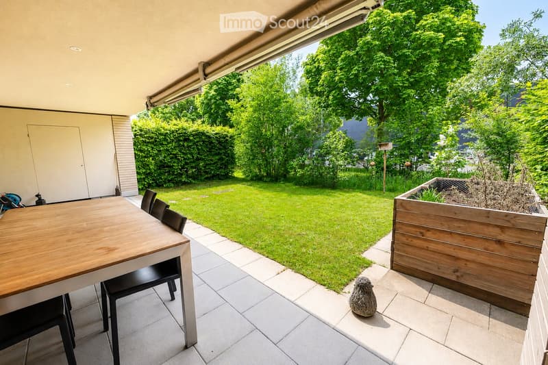 Outdoor patio with a wooden table and black chairs, tiled floor, grassy lawn, wooden planter box, and decorative sculpture.