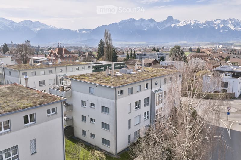Aerial view of a multi-story building with green roofs, surrounded by other buildings and trees, with a view of the mountains in the distance.