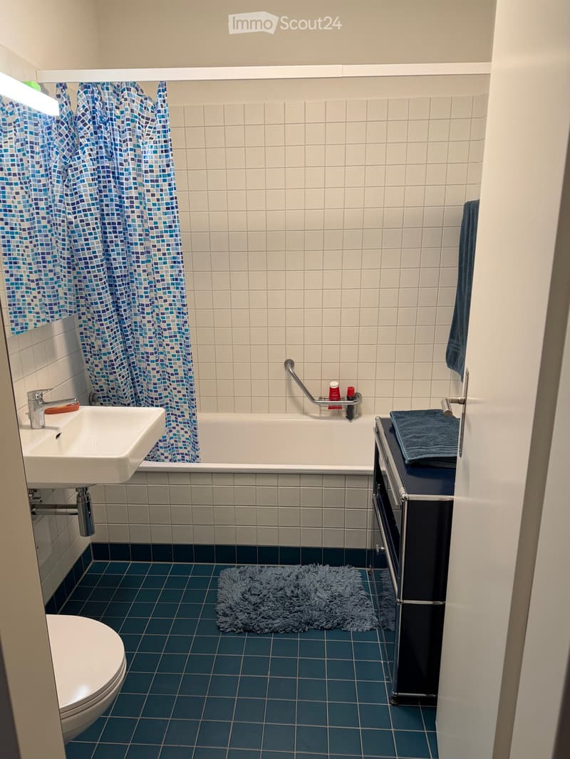 Bathroom with blue and white tiled walls, blue tiled floor, white sink, toilet, bathtub with shower curtain, towel, and a small rug.