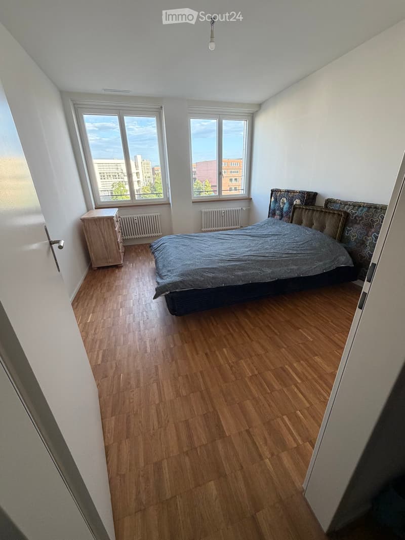 Bright bedroom with wooden flooring, large windows offering a city view, a neatly made bed with patterned bedding, a wooden nightstand, and radiators beneath the windows.