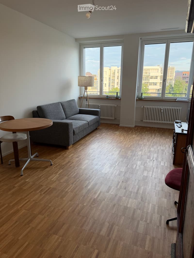 Bright living room with wooden flooring, large windows, grey sofa, round wooden table, and a desk with a chair.