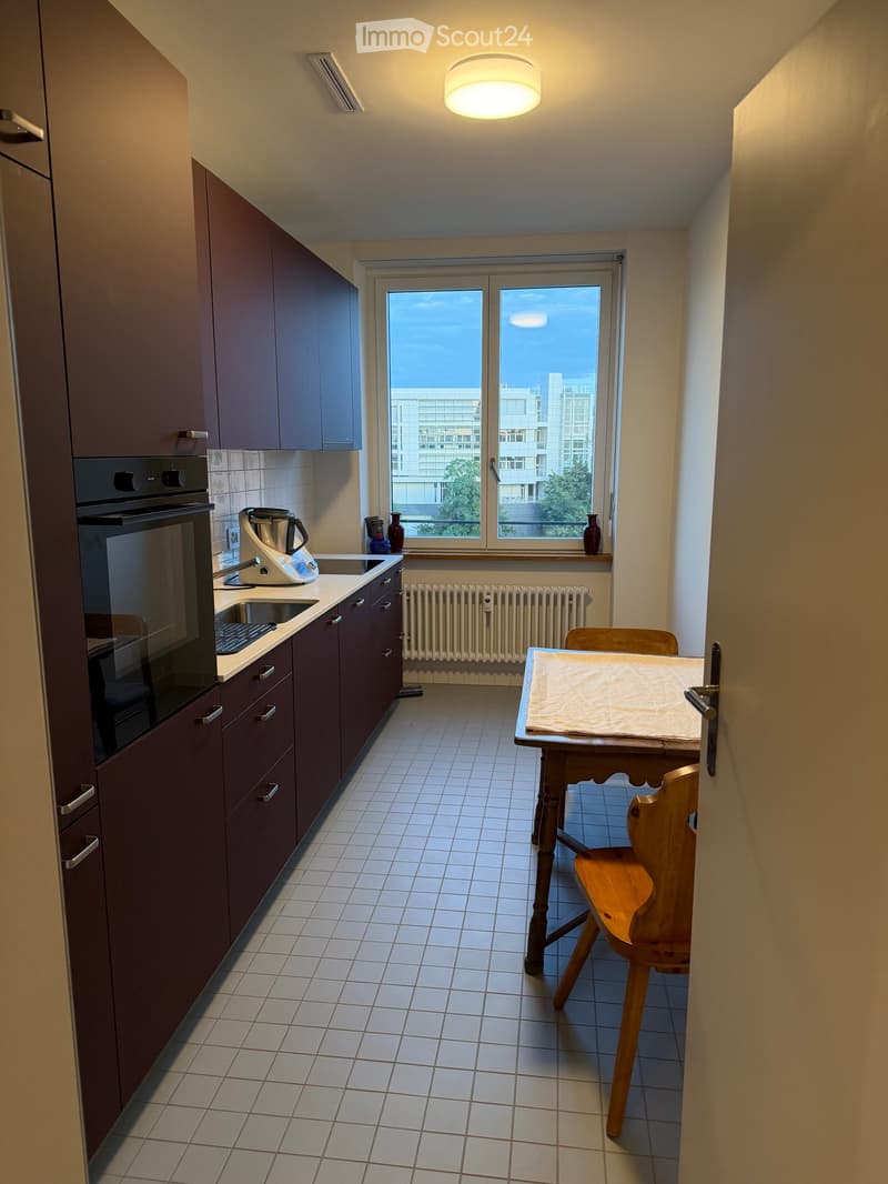 Modern kitchen with dark brown cabinets, white tiled floor, and a window overlooking a cityscape. Includes an oven, sink, table, chair, and decorative vases.