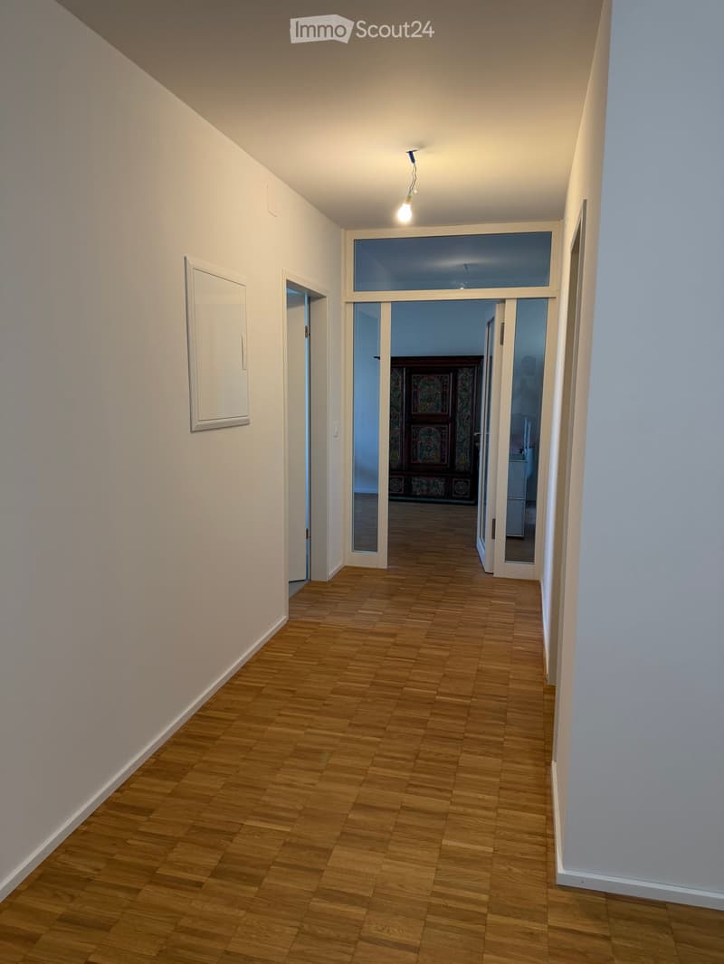 Empty hallway with wooden flooring, white walls, and a single light fixture. A doorway leads to another room with visible furniture and decor.