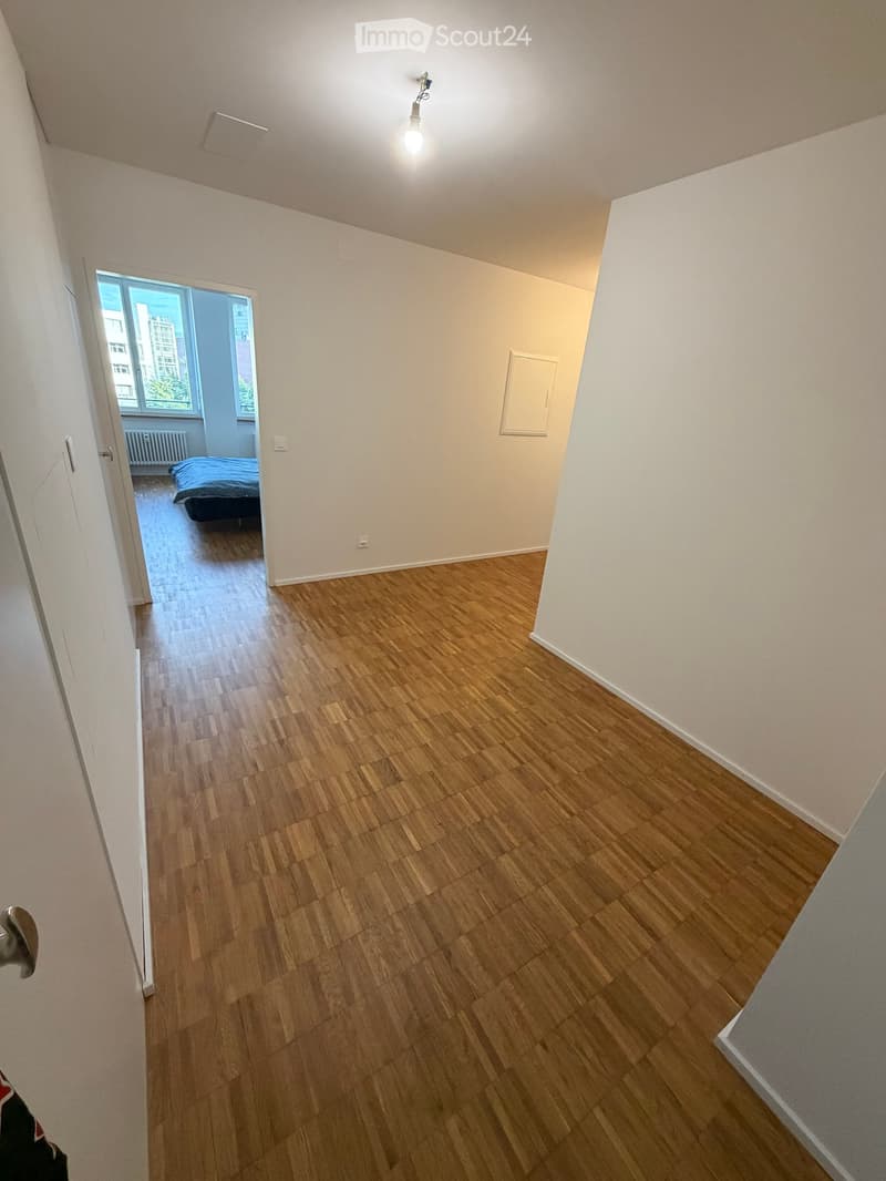 Empty hallway with wooden flooring, white walls, and a visible bedroom with a bed and window in the background.