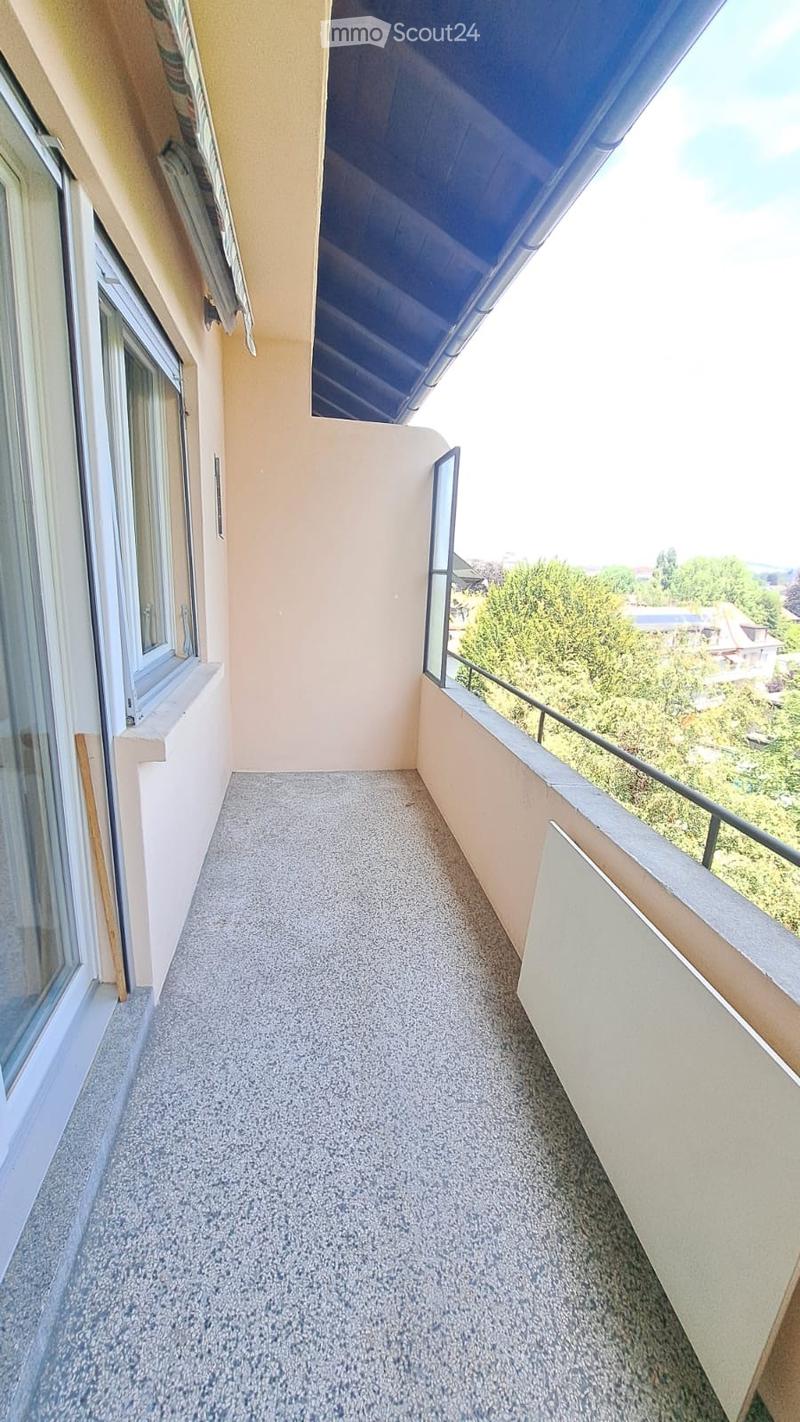 Balcony with grey tiled floor, beige walls, and metal railing, overlooking greenery and distant buildings.