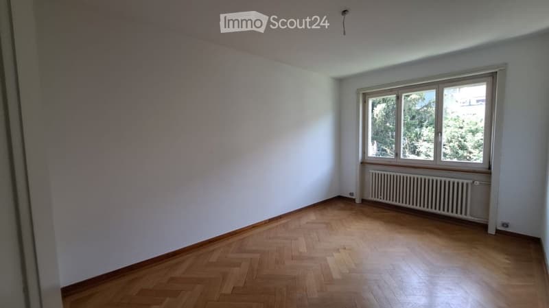Empty room with wooden parquet flooring, white walls, and a large window with a radiator below it.