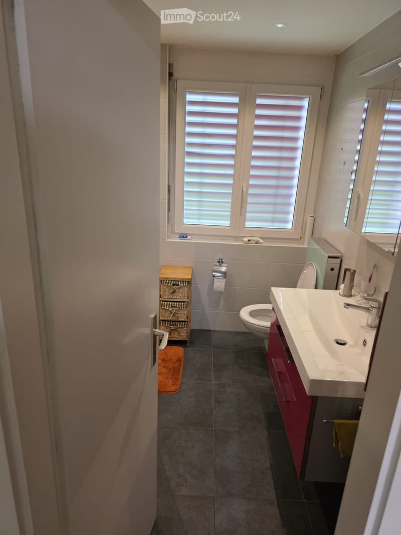 Modern bathroom with a white sink, toilet, mirror, and red cabinet. Features a wicker basket on the floor and a window with blinds.
