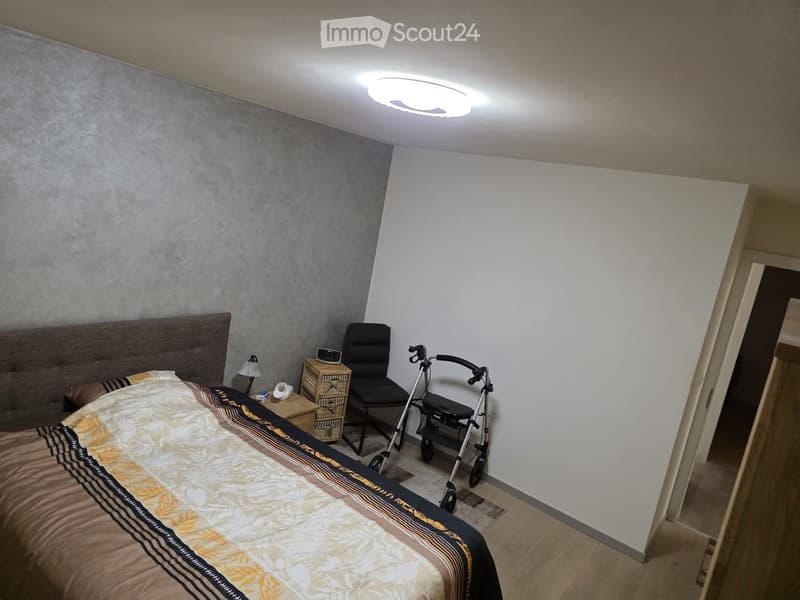 Bedroom with a large bed, nightstand, chair, walker, and lamp. The walls are painted in two tones, and the floor is wooden.