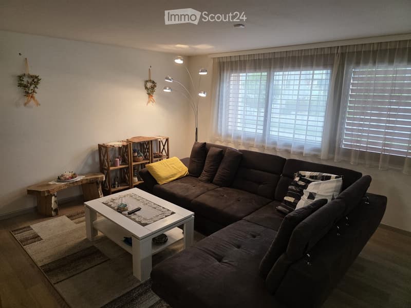 Modern living room with a dark brown sectional sofa, white coffee table, floor lamp, wooden shelf, and decorative wreaths on the wall.