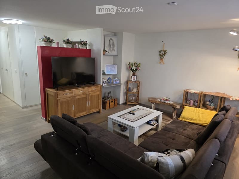 Modern living room with a dark brown sectional sofa, white coffee table, wooden cabinet with TV, and decorative shelves. The room features a mix of wooden and white furniture, with a red accent wall and several decorative items including vases and picture