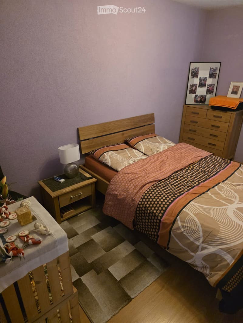 A cozy bedroom with a wooden bed, patterned bedding, a nightstand with a lamp, a small table with decorative items, and a dresser with a picture frame on top.