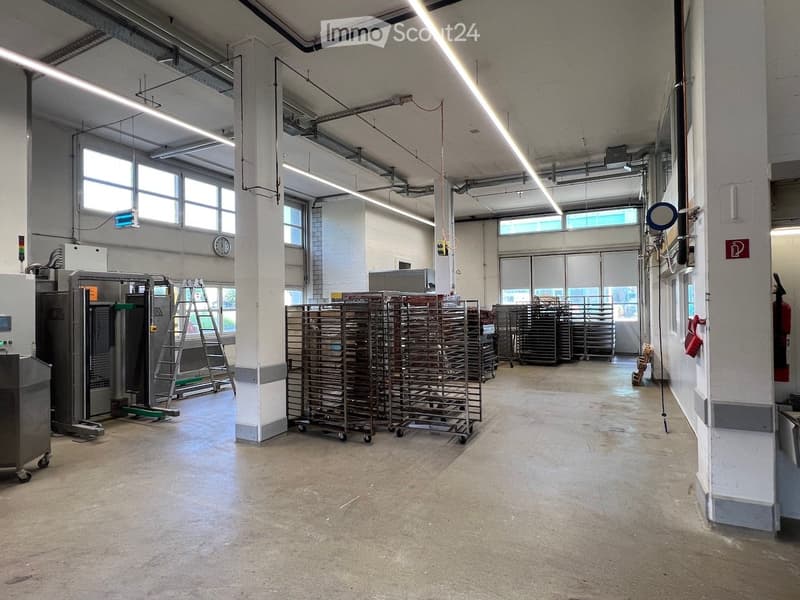 An empty room with concrete floor, large windows, multiple racks, various machines and equipment, 2 ladders, and a clock mounted on the wall.